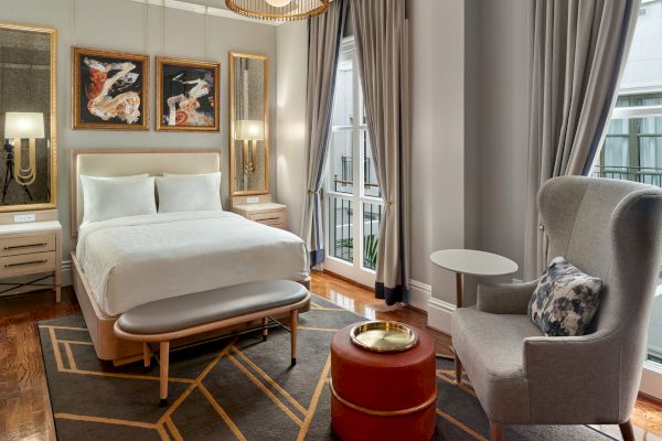 A modern bedroom features a white bed, artworks, bedside tables, armchair, red ottoman, and large windows with curtains, all in a neutral palette.