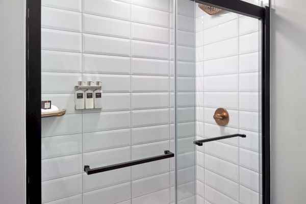 The image shows a modern shower with white tiled walls, a glass door with black handles, and a brass showerhead, along with toiletry bottles.