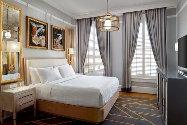 A modern bedroom with a bed, bedside table, artwork on the wall, large windows with curtains, a TV, and contemporary decor.