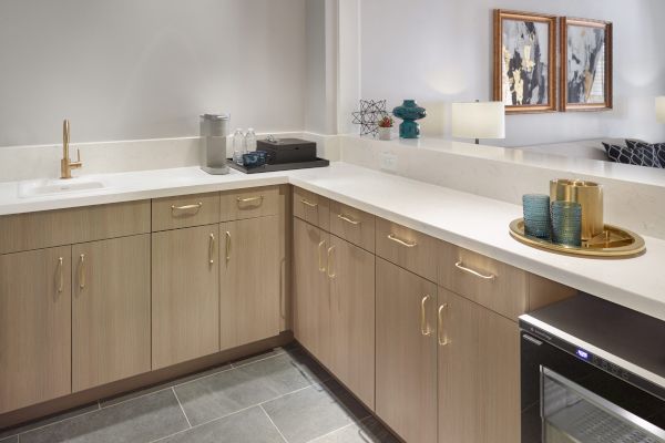 A modern kitchen corner with light wood cabinets, a sink, a coffee maker, a tray with cups, a mini fridge, and wall art in the background.