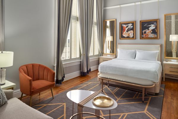 A modern bedroom with a bed, sofa, orange chair, two nightstands, three wall paintings, tall curtains, and a coffee table with a gold tray on a rug.