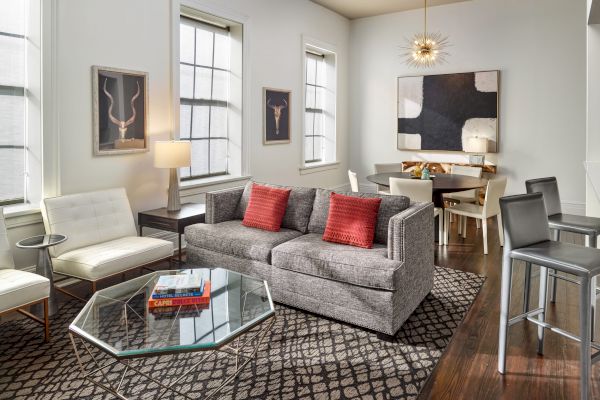 A modern living and dining area features a gray sofa with red pillows, a patterned rug, and abstract wall art. The space is filled with natural light.