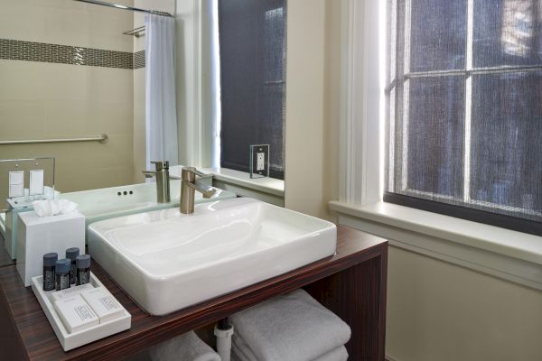 A modern bathroom with a large sink, faucet, toiletries, towels on a wooden shelf, and a window with a dark shade.