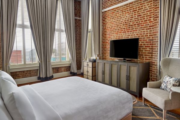 A cozy room with exposed brick walls, large windows with curtains, a bed, an armchair, a TV on a wooden cabinet, and a small side table.