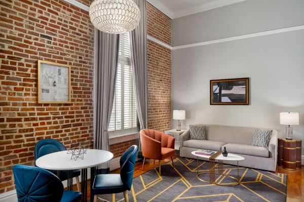 A modern living room with exposed brick walls, a gray sofa, coffee table, blue and orange chairs, and chandelier, plus a dining table and chairs.