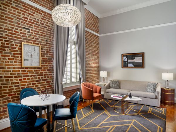 A modern living room with a brick accent wall, gray sofa, blue and orange chairs, a round dining table, a geometric rug, and large windows with curtains.