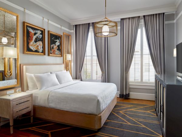 A modern bedroom with a large bed, side table, artwork on the wall, large windows with curtains, a TV, and stylish lighting fixtures.