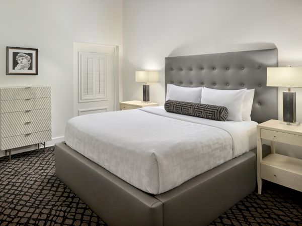 A modern bedroom with a large bed, gray tufted headboard, bedside tables with lamps, a dresser, and framed picture on the wall, featuring a neutral palette.