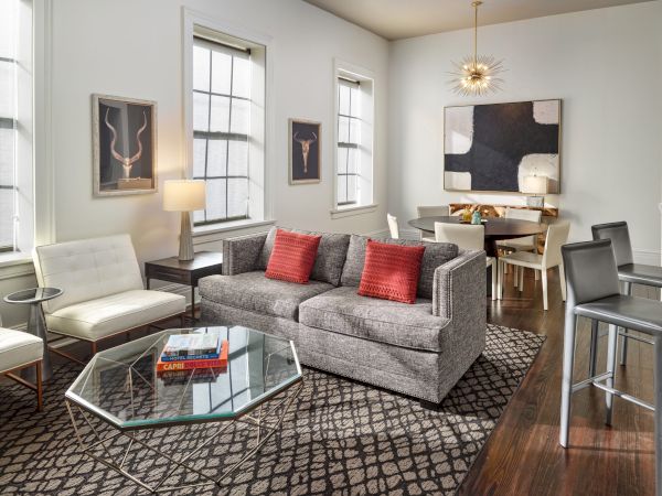 A modern living and dining area with a gray sofa, two white chairs, a glass coffee table with books, dining table, high chairs, and contemporary decor.