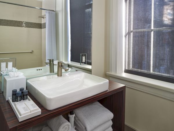 This is a modern bathroom with a rectangular sink on a wooden vanity, toiletries, towels, a mirror above, and a window with black blinds.