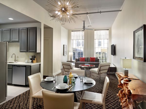 A modern living room with a kitchen, round dining table, seating area, and unique ceiling light fixture, large windows, and a TV on the wall.
