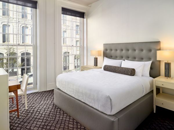 The image shows a stylish bedroom with a king-sized bed, two bedside tables with lamps, and large windows offering a view of a building outside.