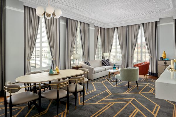 A modern, elegant living room with a dining area, featuring a large patterned rug, multiple windows with drapes, and contemporary furniture.