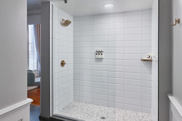 This image shows a clean, white-tiled walk-in shower with a glass door, bronze fixtures, and shampoo/soap dispensers on the wall.