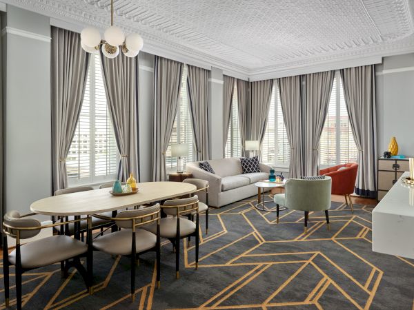 A modern living and dining room with large windows, gray curtains, stylish furniture, a geometric-patterned rug, and decorative items on a console table.