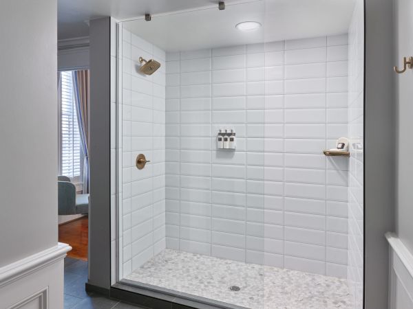 A modern shower with white tile walls, a built-in shelf holding bottles, a showerhead, and a small bench. Light shines in from an adjacent room.