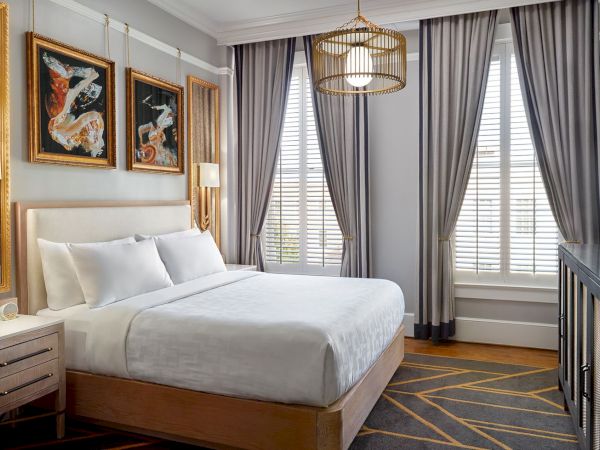 A modern bedroom with a large bed, wooden furniture, art pieces on the wall, two windows with curtains, pendant lights, and a flat-screen TV.