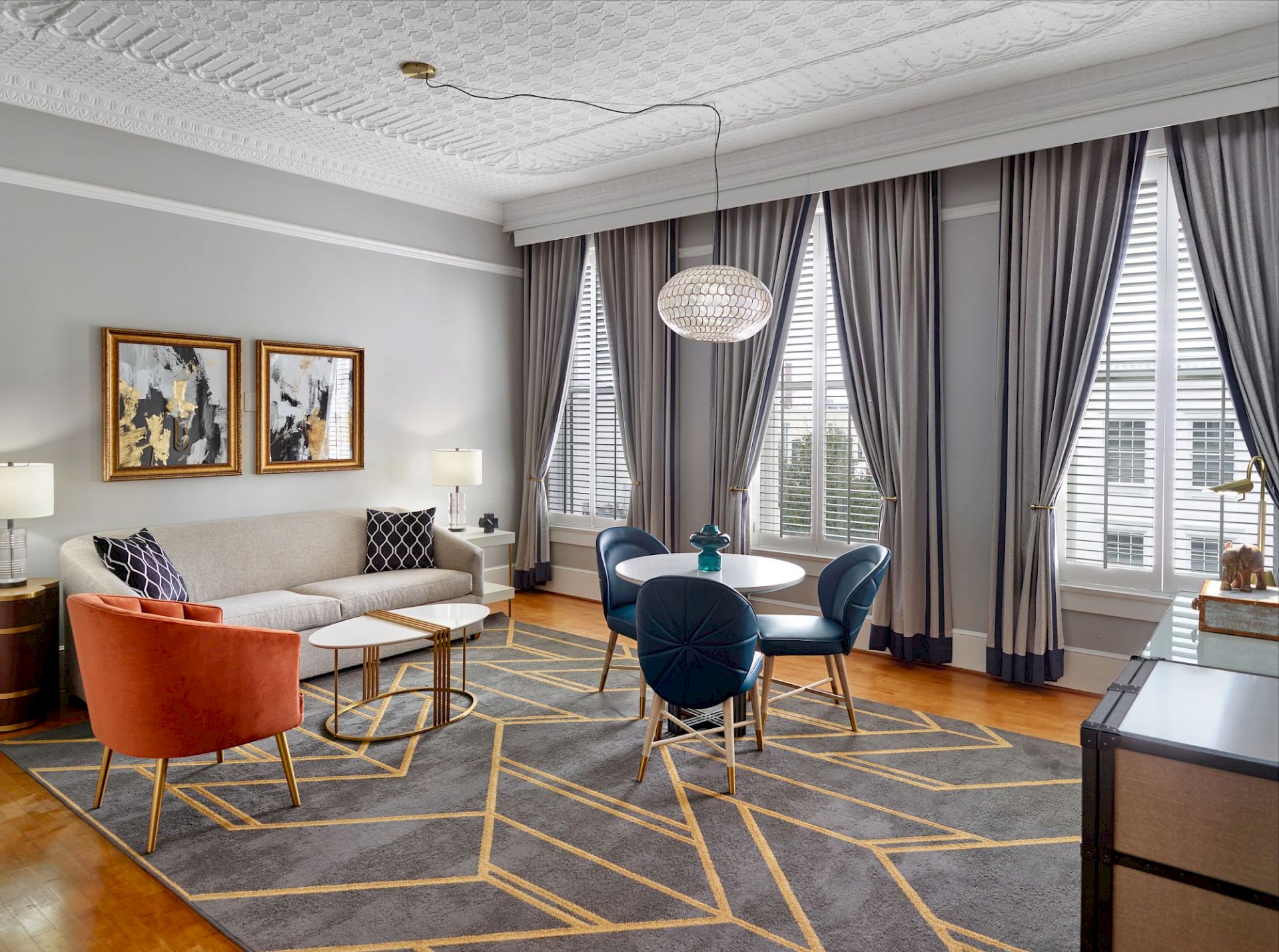 A stylish living room with a sofa, orange armchair, round dining table, blue chairs, modern decor, artwork, a large rug, and tall curtains ending the sentence.