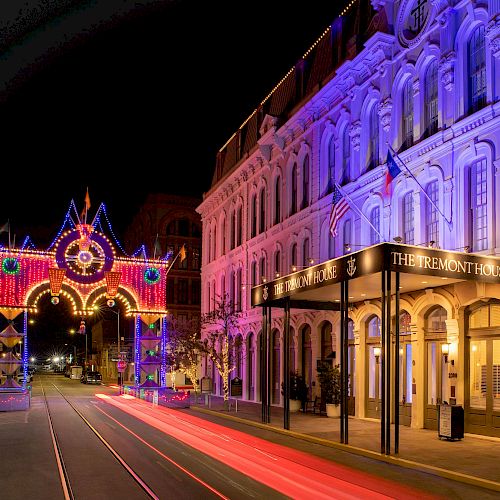 A beautifully lit street at night features an ornate building glowing with purple lights and a brightly illuminated archway in the background, creating a festive atmosphere.