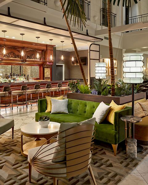 This image shows a stylish, modern lounge with seating areas, a bar, and contemporary decor. Green velvet sofa, chairs, and palm trees are visible.