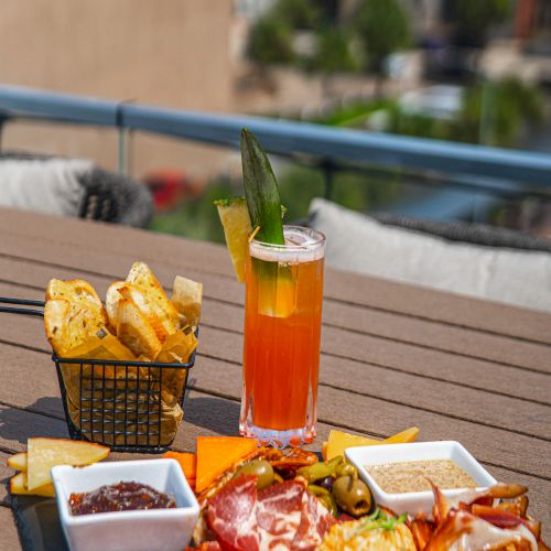 An outdoor patio table with an orange drink, bread, cheese, and olives, featuring a green garnish spear in the glass.