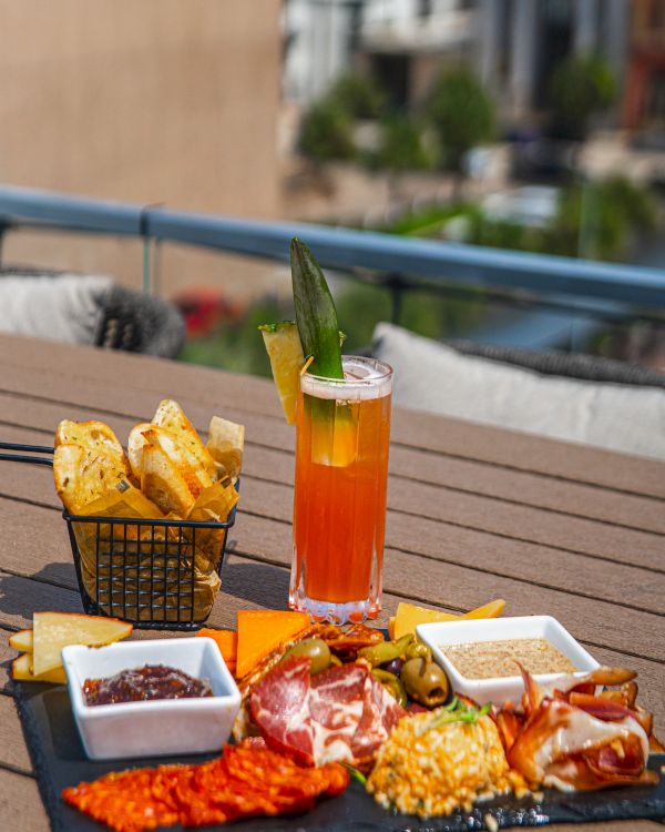 An outdoor patio table with an orange drink, bread, cheese, and olives, featuring a green garnish spear in the glass.
