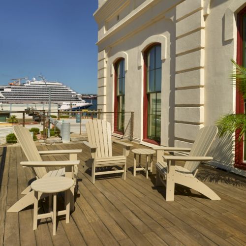 Sunlit wooden deck with beige outdoor furniture, lounge chairs, small tables, a potted palm, beside a light-colored building; a cruise ship seen at harbor.