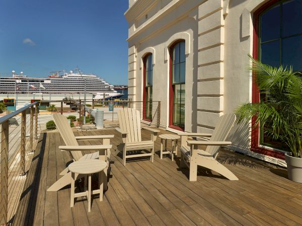 Sunlit hotel balcony with white chairs, palm leaves, and a docked cruise ship in the background.