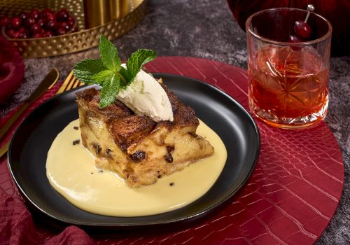 A dessert served on a black plate with custard, topped with ice cream and mint; accompanied by a drink in a glass on a red mat.