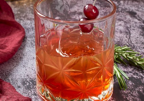 A cocktail served in a decorative glass with ice, garnished with two cherries on a pick. Cranberries and foliage are nearby on the table.