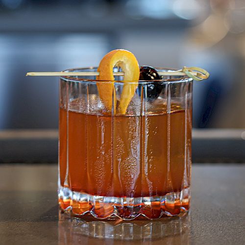 A glass of cocktail with an orange twist and a cherry on a skewer, served in a crystal tumbler.