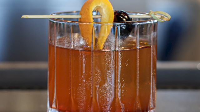 A glass of cocktail with an orange twist and a cherry on a skewer, served in a crystal tumbler.