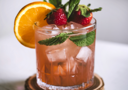 A short, fancy cocktail on a wooden coaster, garnished with a strawberry, mint, and an orange slice.