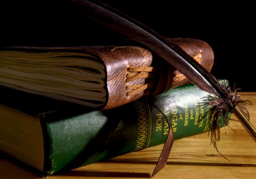 A stack of books with a leather cover and a green spine, topped by a feather quill pen resting on top of the books.