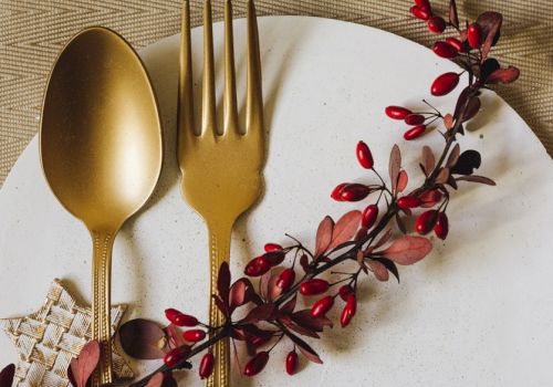 A elegant table setting with gold-toned spoon and fork laid on a white plate, decorated by a red-berry sprig and a woven napkin edge.