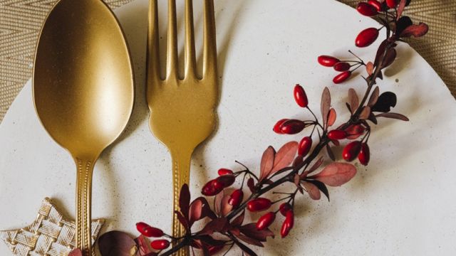 A elegant table setting with gold-toned spoon and fork laid on a white plate, decorated by a red-berry sprig and a woven napkin edge.