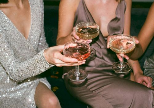 Three women in sparkly and satin dresses clink stemmed glasses of wine or champagne at a party.