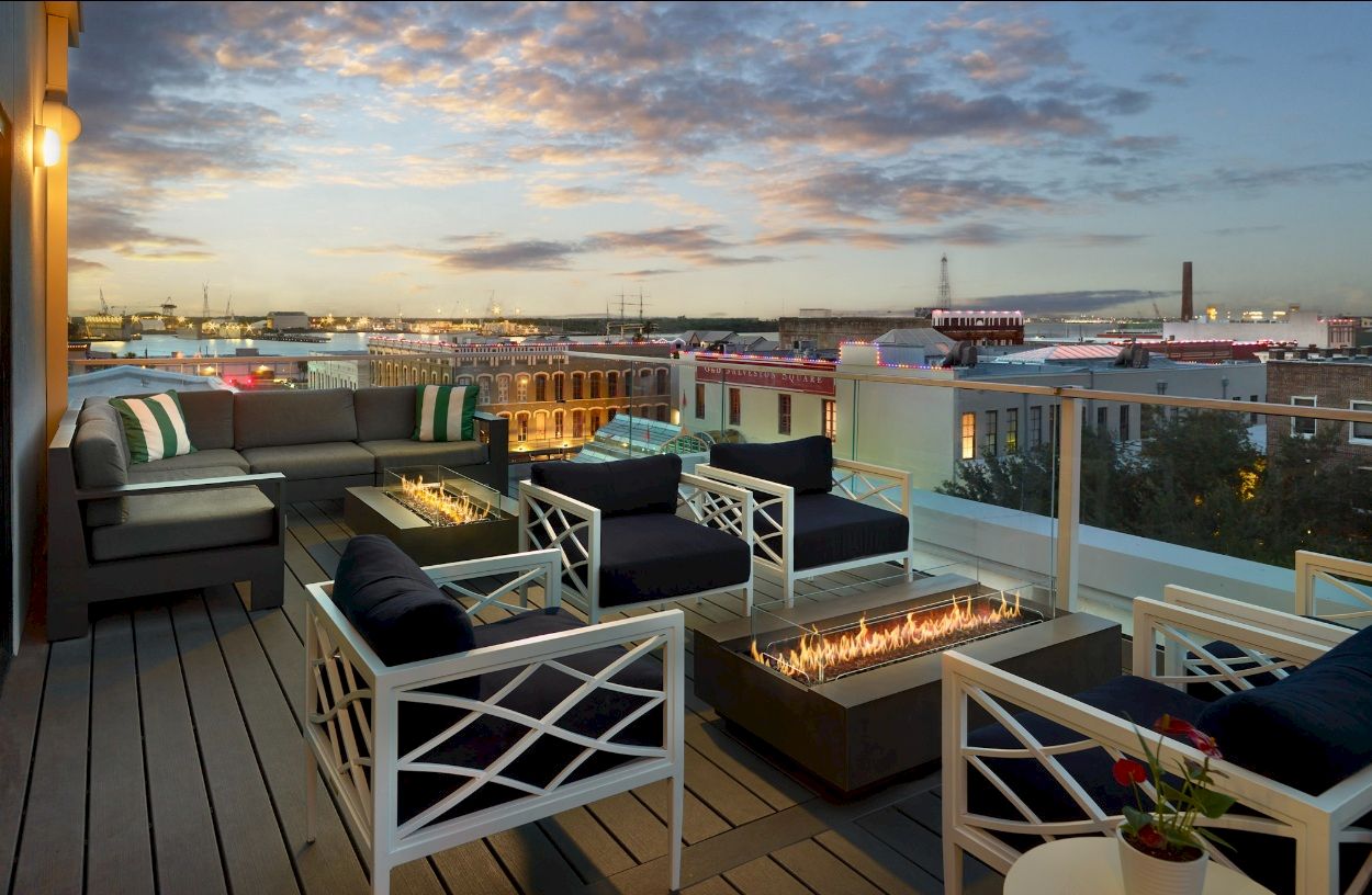 Rooftop terrace with lounge seating and a lit fire pit overlooking a harbor and docks at sunset.