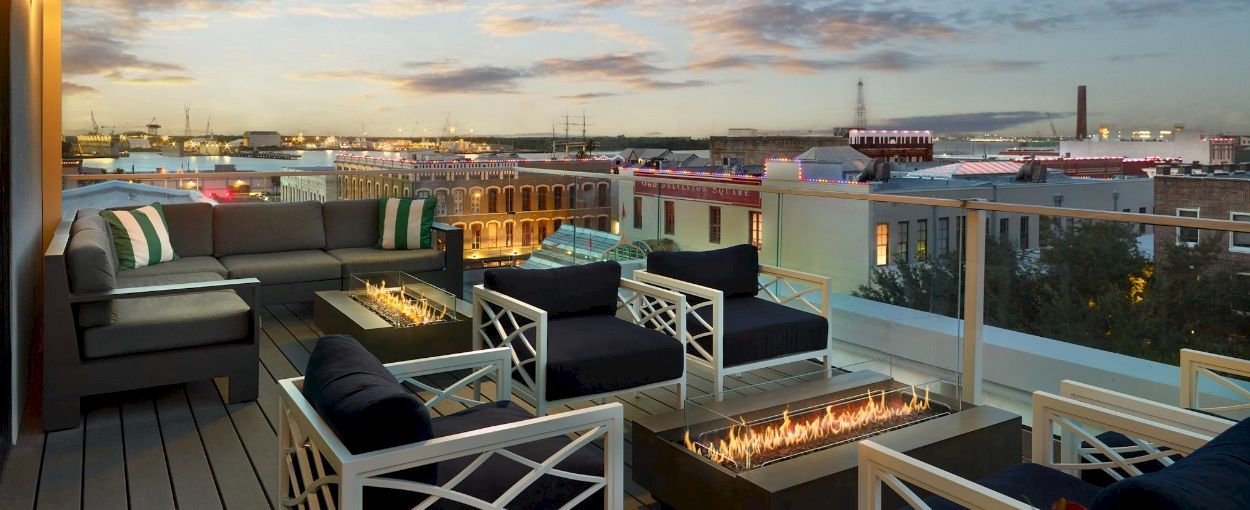 Rooftop terrace with lounge seating and a lit fire pit overlooking a harbor and docks at sunset.