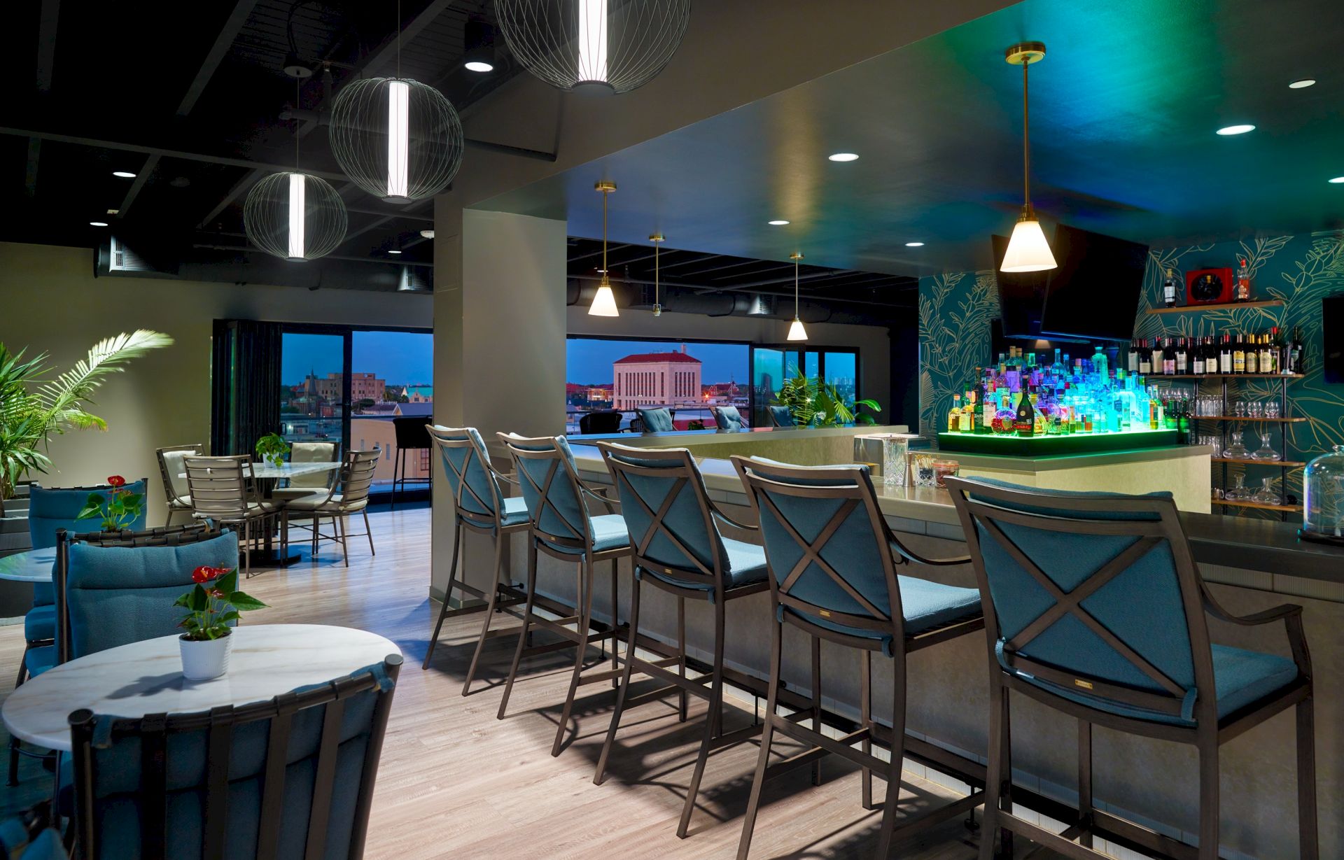A modern bar lounge with a glowing green bottle display, pendant lamps, lounge chairs, and large windows overlooking the city at dusk.