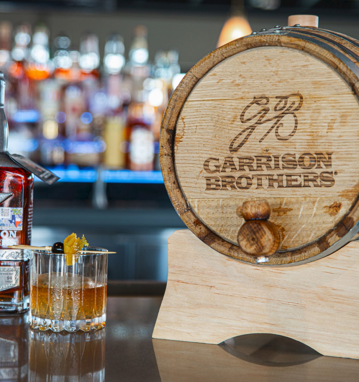 A Garrison Brothers bourbon bottle beside a branded wooden barrel in a bar setting.