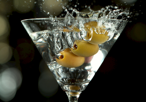 A martini glass filled with amber liquid is caught mid-splash, with bubbles and droplets bursting upward.