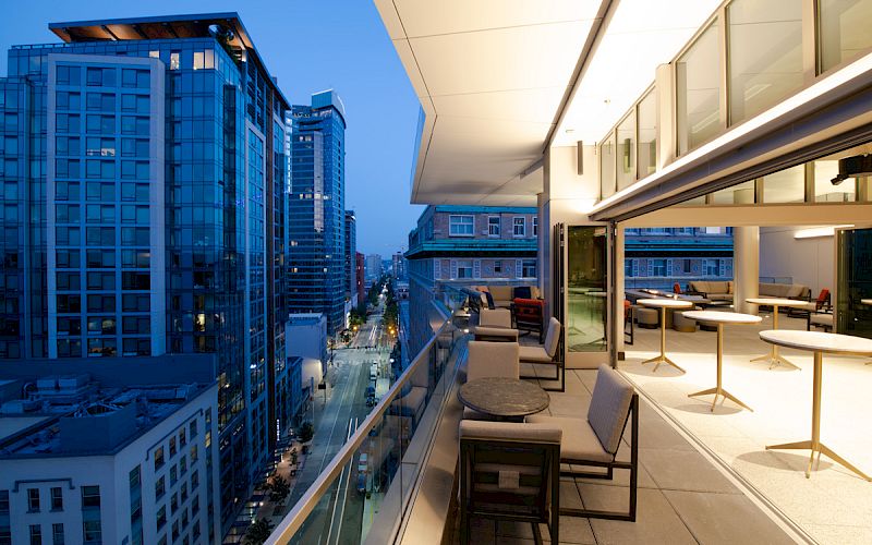 A cityscape at dusk, with a view of modern buildings and a stylish outdoor terrace featuring tables and chairs extending from an indoor space. The end.