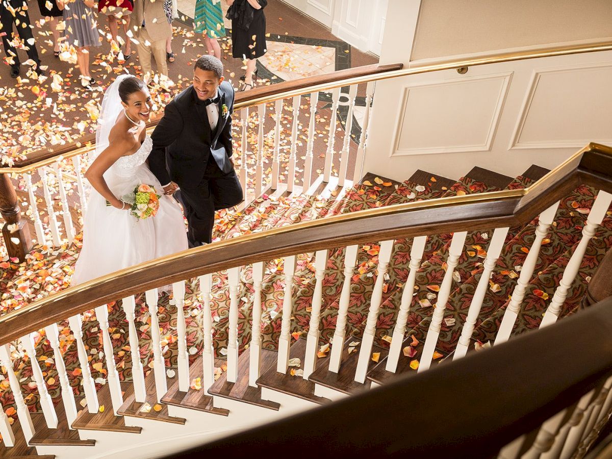 A bride and groom are walking up a staircase adorned with floral decorations, surrounded by people celebrating.