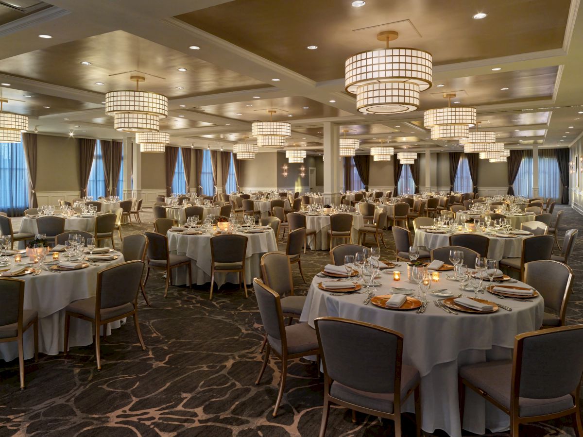 A large banquet hall with round tables set for an event, featuring elegant chandeliers and floor-to-ceiling windows with curtains.