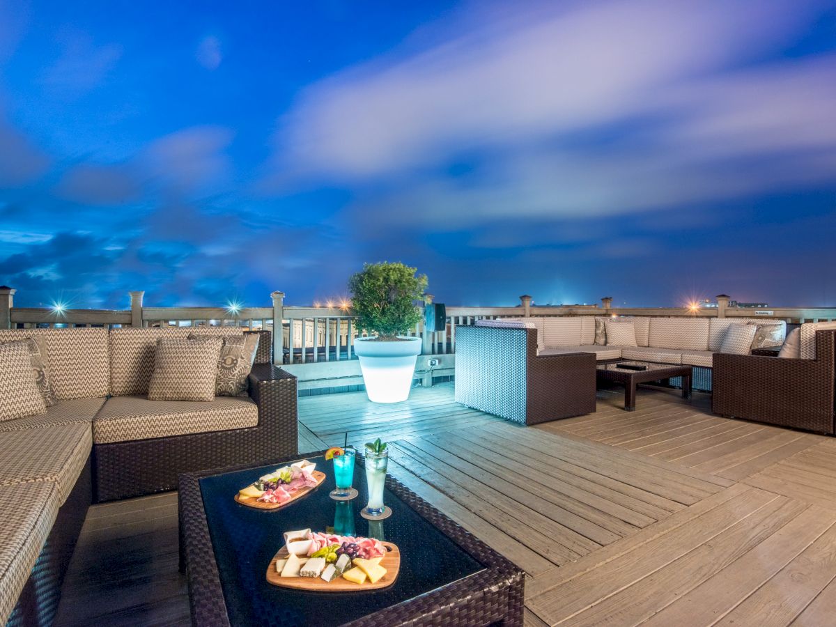 A rooftop terrace with wicker furniture, snacks on a table, soft lighting, and a beautiful evening sky in the background.